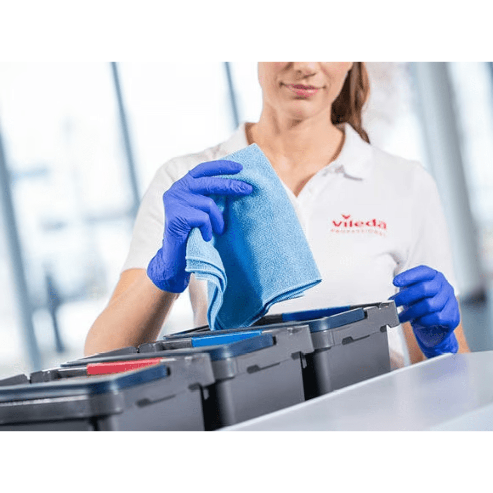 A person wearing a white shirt and blue gloves is holding a blue Vileda Professional r-MicroTuff Swift microfiber cloth from Vileda GmbH over a row of containers with lids of different colors; the scene illustrates the efficient cleaning process using the four-color organization system.