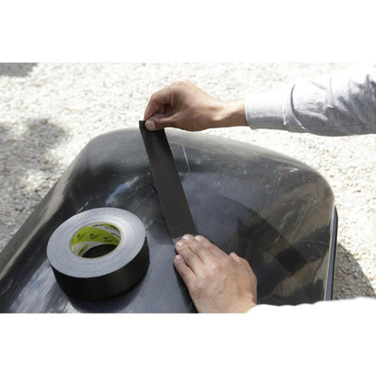 A person in a long-sleeved shirt applies the Scotch® Extremium™ Ultra High Performance Tape DT17 (10 m x 24 mm) by 3M Deutschland GmbH to a smooth, dark surface outdoors to demonstrate its use for extreme demands, with the tape roll nearby.