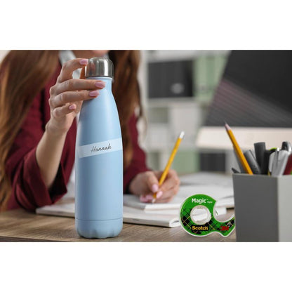 A person sits at a desk with a light blue "Hannah" water bottle. On the desk are a notebook, pencils, pens in a cup, and the Scotch® Magic™ Invisible Tape (19 mm x 25 mm) with dispenser (+5 m free) by 3M Deutschland GmbH. Behind are trays and folders.