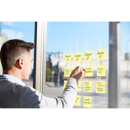 A person arranges Post-it® Super Sticky Notes (47.6 x 47.6 mm, 90 sheets/block, 100% PEFC) from 3M Deutschland GmbH on a window to outline project phases - presentations, status checks, and completion - while sunlight streams in.
