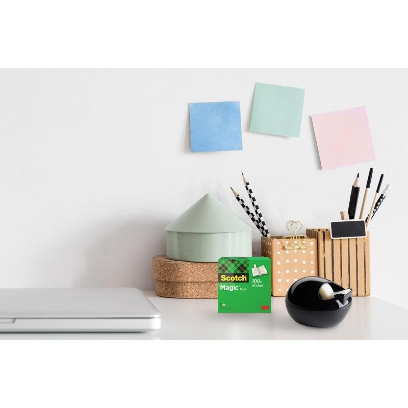 A desk with a closed laptop, 3M Scotch™ Magic Tape, a black Scotch™ Karim Rashid tape dispenser from 3M Deutschland GmbH, cups with pens and pencils, stacked round boxes, and three pastel sticky notes on a white wall.