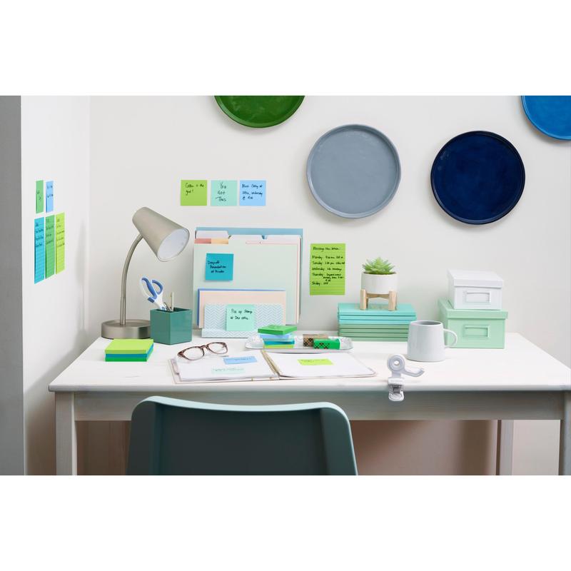 A tidy white desk with office supplies, a lamp, file folders, a plant, glasses, boxes, and 3M Post-it® Super Sticky Z-Notes (Boost Collection). Colorful plates hang above it, and Post-it® notes are arranged on the wall and on the desk.