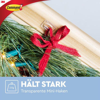 A Command™ Mini Hook, transparent (3M Deutschland GmbH), is applied residue-free on wood and holds a pine branch bundle with a red ribbon (up to 225g). Text: "HOLDS STRONG Transparent Mini Hooks".