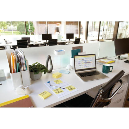 A modern office with a laptop, 3M Post-it® Notes (yellow, 76x76 mm, 100 sheets/block), a cup, documents, plants, and accessories on a white desk. In the background, more desks and large windows can be seen.