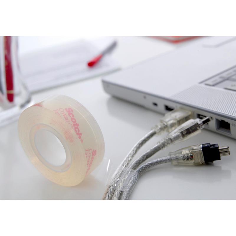 A roll of Scotch® Crystal Tape, transparent (19 mm x 33 m) by 3M Deutschland GmbH lies on a white desk next to a laptop and cables; in the background, papers and a pen are visible.