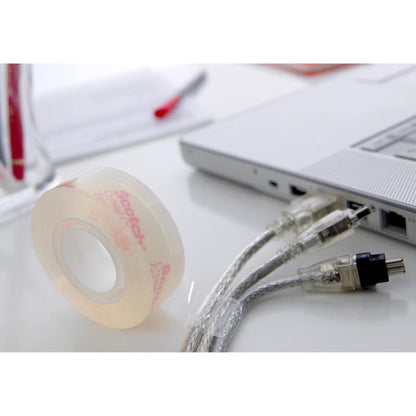 A roll of Scotch® Crystal Tape, transparent (19 mm x 33 m) by 3M Deutschland GmbH lies on a white desk next to a laptop and cables; in the background, papers and a pen are visible.