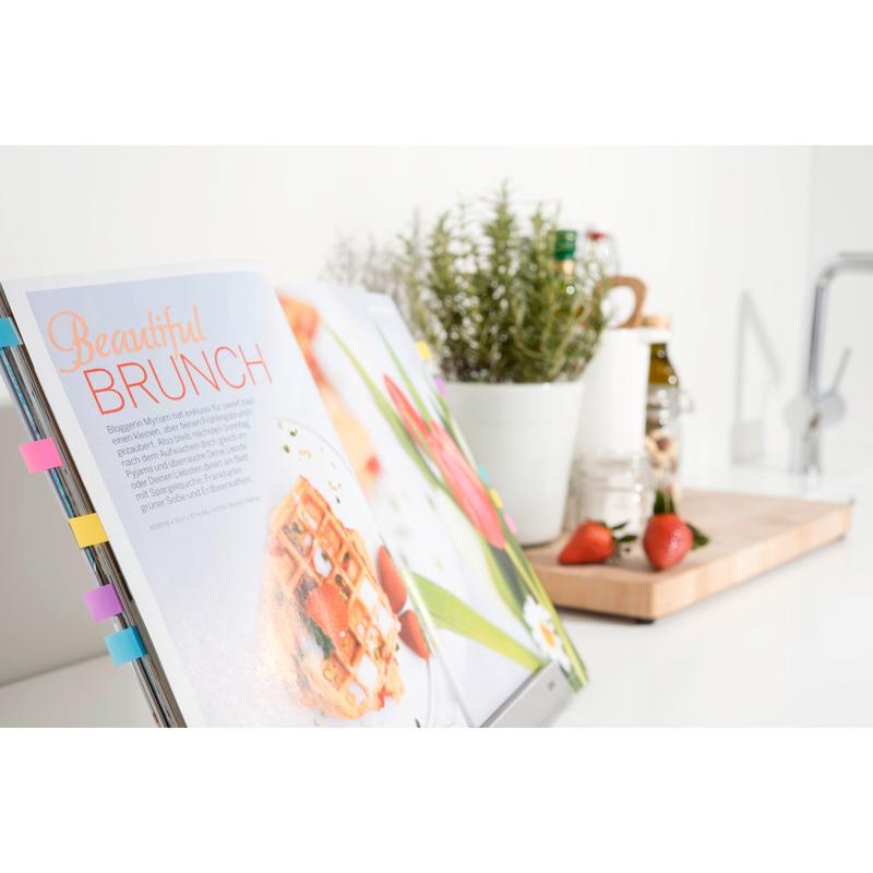 An open cookbook on a stand is labeled with colorful 3M Deutschland GmbH Post-it® Index Flags (Mini, various colors) and Post-it® Index Arrows (Blue and Yellow), with herbs, bottles, strawberries, and a cutting board next to it.