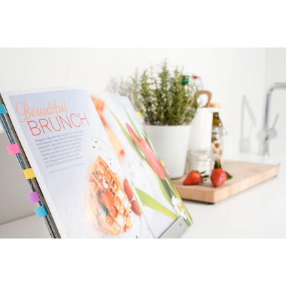 An open cookbook on a stand is labeled with colorful 3M Deutschland GmbH Post-it® Index Flags (Mini, various colors) and Post-it® Index Arrows (Blue and Yellow), with herbs, bottles, strawberries, and a cutting board next to it.
