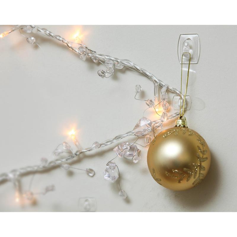 A golden Christmas ornament hangs on a Command™ Mini Hook, transparent (17006CLRFGN) by 3M Deutschland GmbH, on a white wall next to glowing white Christmas lights - perfect storage without drilling!