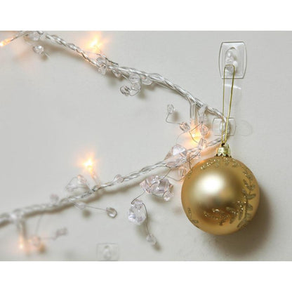 A golden Christmas ornament hangs on a Command™ Mini Hook, transparent (17006CLRFGN) by 3M Deutschland GmbH, on a white wall next to glowing white Christmas lights - perfect storage without drilling!