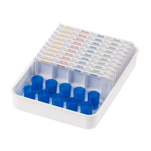 A white Melipul dispenser/cup tray from Helmut Schwarz GmbH contains several transparent pill dispensers, part of a medication organization system, labeled with German words for the time of day: "Morning", "Noon", "Evening", "Night". Each reusable dispenser features slots for pills and blue lids on one side.