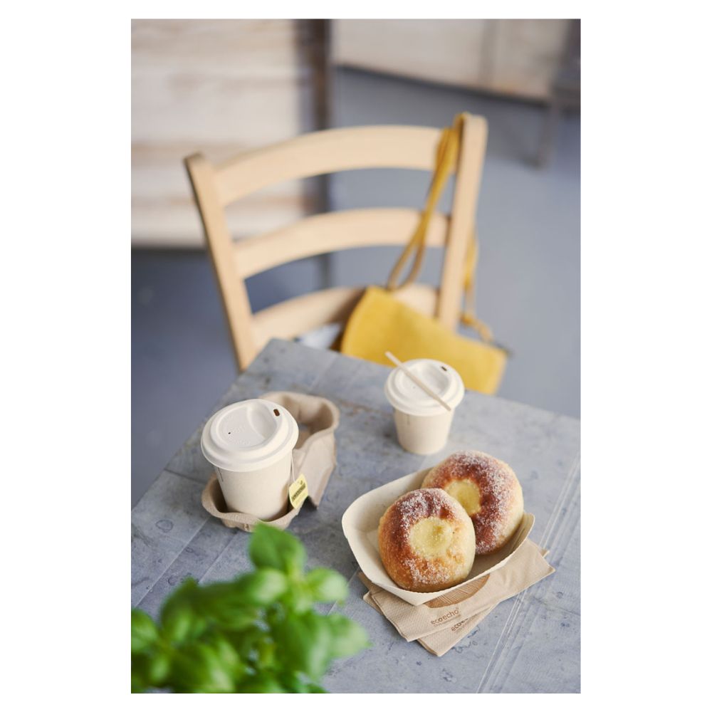 On a table, there are two pastries dusted with sugar on a tray and two Duni coffee cups 'Sweet Printed Bagasse' with lids from Duni GmbH. A yellow handbag hangs on a wooden chair, while a partially visible green plant in the foreground highlights the subtle theme of sustainability.