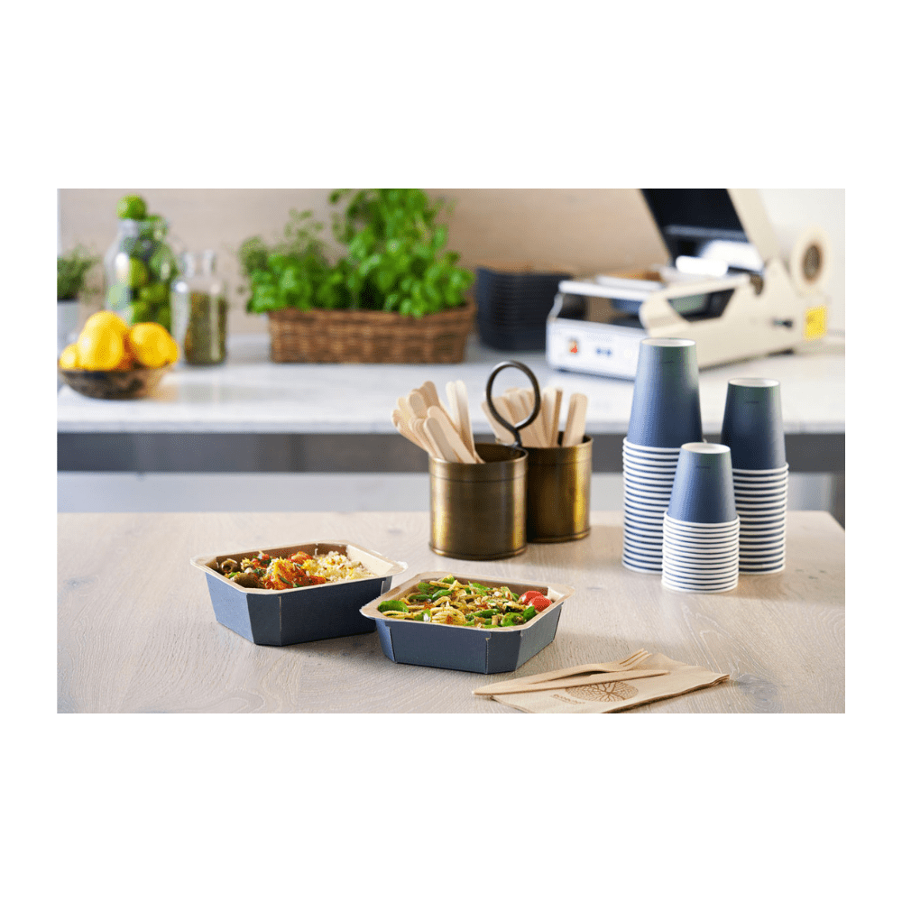 On the table are two take-away bowls, flanked by stacked Duni Urban Coffee Cups PAP/PLA from Duni GmbH, wooden cutlery in a holder, and napkins. Lemons, glasses, and herbs in the background contribute to the sustainable ambiance of this food preparation area.
